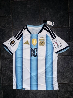 Argentina Messi matchtröja Adidas - Säljer en officiell Argentina matchtröja från Adidas med Messi och nummer 10 på ryggen. Tröjan har klassiska blå och vita ränder, guldiga detaljer, FIFA- och AFA-emblem samt svarta ränder på axlarna. Korta ärmar och rund hals. Perfekt för fotbollsfans!