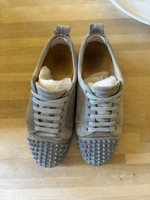 Loubs me spikar - Säljer ett par grå sneakers från Christian Louboutin i mocka med grå spikar på tån och klassisk snörning. Skorna har en chunky sula och en stilren look som sticker ut. Perfekta för dig som vill ha något unikt till vardagslooken. Har lite skador där bak och där fram, har även tappat en spik.