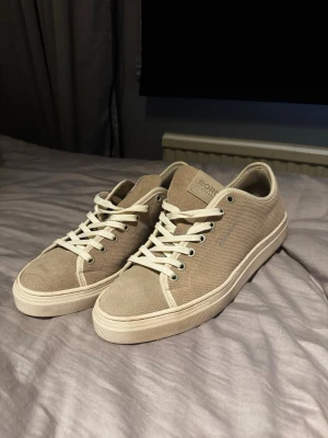 Beige canvas sneakers med vita detaljer - Snygga beige sneakers i canvas med vita snören och vit sula. Klassisk låg modell med enkel design som passar till det mesta. Skorna har metallöljetter och diskret logga på plösen. Perfekta för dig som gillar stilrena och bekväma sneakers. Näst intill nyskick 