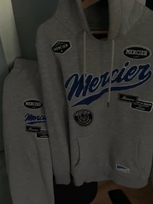 Grå tracksuit från Mercier med tryck - Säljer en grå tracksuit från Mercier med flera svarta och vita patches samt ett stort blått Mercier-tryck. Hoodien är storlek M medan byxorna är storlek L. Hoodien har huva med dragsko och en stor magficka. Perfekt för dig som gillar streetwear och vill sticka ut lite extra. Pris kan diskuteras.