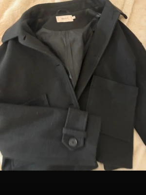 Svart klassisk ullkappa - Stilren svart ullkappa med krage och knäppning framtill. Kappan har raka ärmar med dekorativ knappdetalj vid ärmslutet och stora fickor. Perfekt för en trendig och tidlös look under kyliga dagar.