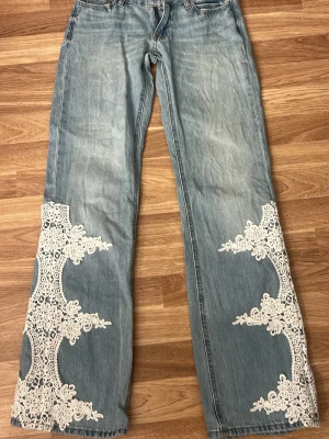 Ljusblå jeans med spetsdetaljer - Kostar 499 i butik, använda typ 2 gånger, Unika ljusblå jeans med breda ben och stora vita spetsapplikationer på både fram- och baksidan av benen. Jeansen har klassisk femficksmodell och är tillverkade i denim. Perfekta för dig som vill sticka ut med en kreativ och bohemisk stil.