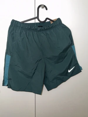 Mörkgröna träningsshorts från Nike - Snygga mörkgröna träningsshorts från Nike med elastisk midja och vita loggan på vänster ben. Shortsen har ljusare gröna paneler på sidorna och är tillverkade i ett lätt, syntetiskt material som andas. Perfekta för gymmet eller löprundan.