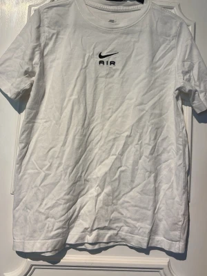 Vit Nike Air t-shirt med tryck - Snygg vit t-shirt från Nike med svart 'AIR' tryck och logga både fram och bak. Klassisk rund hals och korta ärmar. Perfekt till en avslappnad streetwear-look. Tillverkad i mjuk bomull för skön känsla hela dagen.