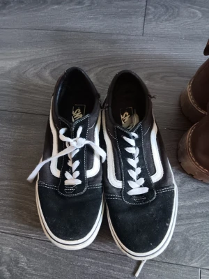 Svarta Vans Old Skool sneakers - Klassiska Vans Old Skool sneakers i svart med vit sula och vit sidostripes. Skorna har lågt skaft, vita snören och är tillverkade i en kombination av mocka och canvas. Perfekta för en avslappnad streetwear-look.