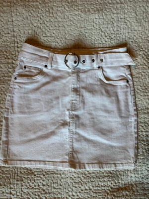 Vit kortkjol med bälte - Snygg vit kortkjol i denim-look med bältesdetalj. Kjolen är stretchig och har fickor fram och bak.