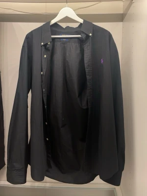 Svart skjorta från Polo Ralph Lauren - Säljer en svart skjorta från Polo Ralph Lauren i slim fit-modell. Skjortan har klassisk krage, knappar framtill och en liten lila logga broderad på bröstet. Perfekt för dig som gillar stilrena och tidlösa plagg.