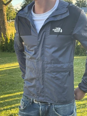 Grå vindjacka från The North Face - Snygg grå vindjacka från The North Face med svarta detaljer på axlar, ärmar och huva. Jackan har dragkedja, tryckknappar, två stora fickor och en justerbar huva. Perfekt för blåsiga dagar och outdoor-äventyr. Finns tecken på användning men är i stort sätt i utmärkt skick.