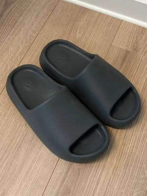 🖤Svarta Adidas Yeezy slides🩶 - Säljer ett par mörkgråa Adidas tofflor i mjukt och flexibelt skummaterial. De har en chunky design med bred rem över foten och öppen tå. Perfekta för att glida runt hemma eller till och från gymmet. Har du storlek 40-41 så passar de dig. Jag har bara använt dem 1 gång ute.