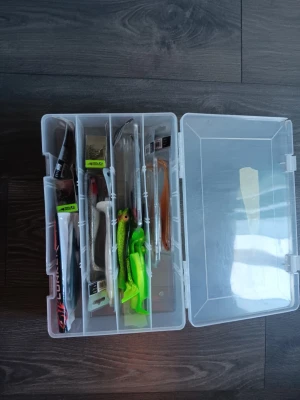 Fiskedrag i plastlåda - En genomskinlig plastlåda fylld med olika fiskedrag i flera färger, bland annat grönt, vitt, brunt och orange. Drag i olika former och storlekar, vissa med fiskliknande utseende. Perfekt för sportfiske och fiskeentusiaster. Nya 