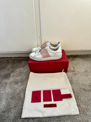🎀 Valentino Garavani Open Sneakers – Stl 37 🎀 - Säljer mina supersnygga och klassiska Valentino Garavani Open Sneakers i vitt läder med det ikoniska ljusrosa bandet i lack samt nitar på hälen. En riktig drömsko som passar till precis allt – från jeans till klänning! • Märke: Valentino Garavani • Modell: Open Sneaker (RW2S0781 TCQ) • Storlek: 37 • Färg: Vit / Ljusrosa (Bianco/W) • Nypris: Ca 6 500 – 7 500 kr  Skorna är använda men har massor av liv kvar att ge! 