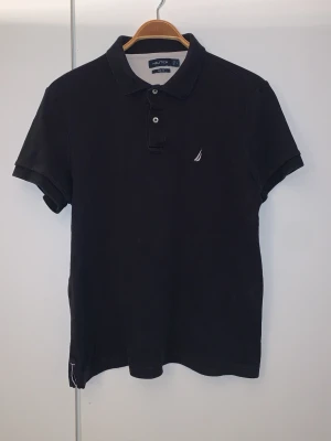Svart pikétröja från Nautica - Klassisk svart pikétröja från Nautica i slim fit-modell. Tröjan har korta ärmar, krage och knappar framtill samt en liten broderad logga på bröstet. Perfekt för en stilren och avslappnad look.