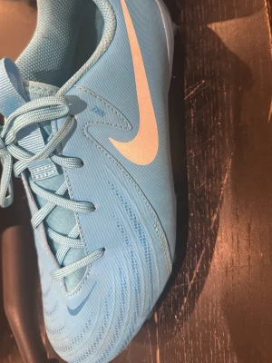 Blå Nike fotbollsskor med snörning - Snygga ljusblå fotbollsskor från Nike med klassisk vit swoosh på sidan. Skorna har snörning och är tillverkade i ett syntetmaterial med textilfoder. Perfekta för dig som vill sticka ut på planen med stilrena detaljer och modern design.