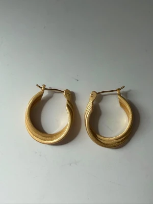 Guldpläterade creoler med vågig design - Snygga örhängen i form av creoler med en unik vågig struktur. De är guldpläterade och har en matt finish som ger ett modernt intryck. Perfekta för dig som gillar stilrena men ändå iögonfallande accessoarer.