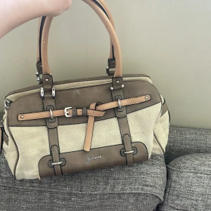 Guess Väska - Äkta Guess väska med neutrala skinndetaljer, den är i använt/gott skick. Crossover bandet har släppt, går säkert att lämna in för lagning. Kommer med dustbag.