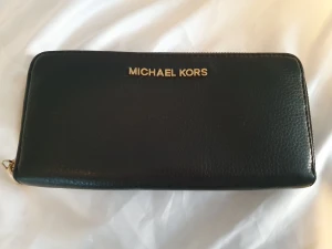 Michael Kors stor plånbok  - Stor plånbok från MK som tyvärr inte kommer till användning. Svart med gulddetaljer. Myntficka med dragkedja och plats för ett flertal kort och andra saker Har ibland även haft mobilen däri.  Höjd: 10.5 cm, Längd: 21 cm Vidd: 2.5 cm 