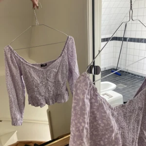 Lila gripas tröja med puffärm - Lila croppad tröja med puffärmar, vitt blommönster och volanger. Storlek Xs men passar S. 