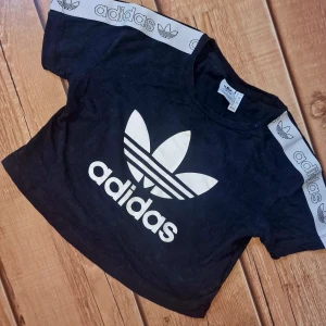 Crop top från ADIDAS - Svart crop-top med stor adidaslogga och ränder längst med ärmarna. Storlek S. Använd ett fåtal gånger, lika fräsch som ny-skick. Äkta adidas.