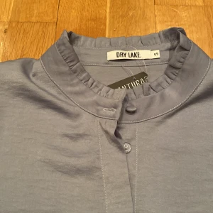 Blus Dry Lake oanvänd - Jättefin duvblå/grå blus från Dry Lake med fina detaljer. Silkigt tyg. Helt oanvänd, lapp kvar. Nypris ca 600 kr. 