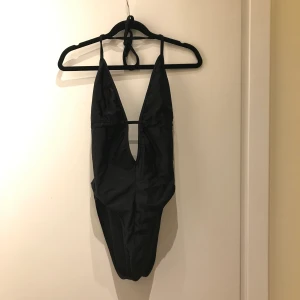 Svart baddräkt  - Svart baddräkt, djup urringning bak och fram. Knyts bakom nacke samt på ryggen. Sparsamt använd 