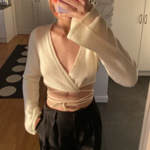 Omlott stickad tröja - Säljer denna fina stickade tröjan från NA-KD. Tröjan är naturvit och v-ringad. Den har långa snören som man kan knyta runt magen bland annat. Storlek S.