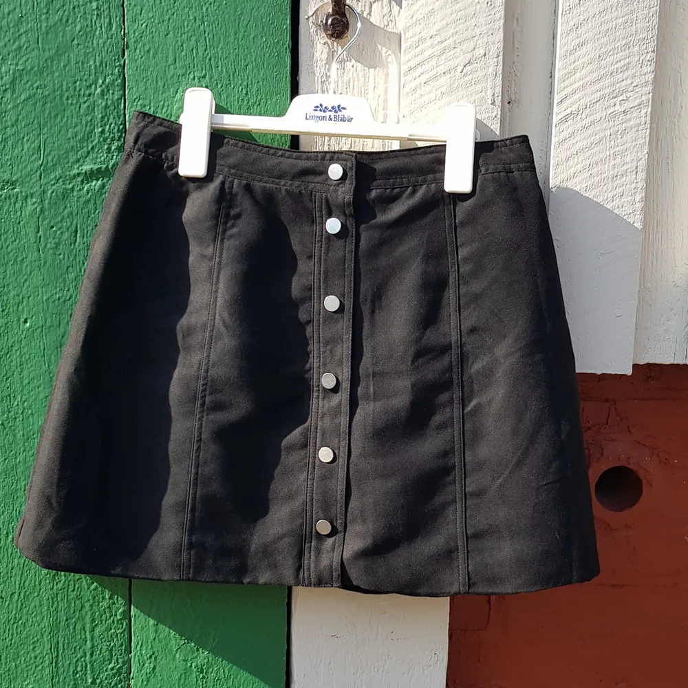 Oanvänd svart, a-linje kjol i storlek 40, tyget känns som fejk mocka och består av 100% polyester. Från H&M Divided. Knappar med knäppning. . Kjolar.