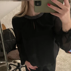 Fin svart blus med detalj - Hej! Säljer nu denna blus då den börjar bli lite liten för mig💗 Har haft den över 1 år men bara använt den 3/4 ggr. Därav är den i nyskick!  Detalj finns runt halsen (se bild 2) + knäppe vid nacken✨