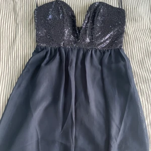 Kort festklänning - Helt oanvänd festklänning. Axelbandslös med djup ringning fram. Glittrig överdel. Köparen betalar frakt