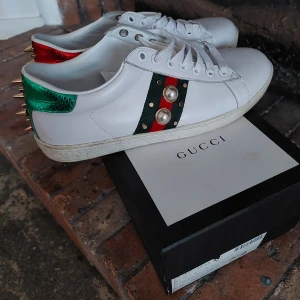 Gucci sneakers - Vita sneakers med nitar och pärlor och röd/grön metallic. Storlek 40, passar mer stl 39-39,5. Sparsamt använda i toppenfint skick! Extra nitar medföljer, ifall någon trillar bort med tiden.