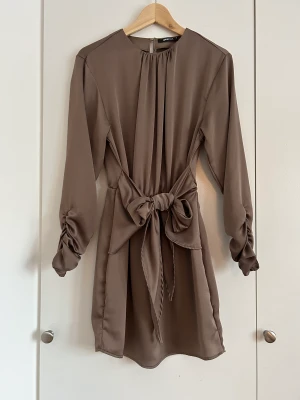 Satinklänning med knyt i midjan och rynkning vid ärmarna - Jättefin satinklänning med knytning i midjan och jättesnygga rynkningar vid ärmslutet.  Jättebra skick, använd vid fåtal tillfällen.