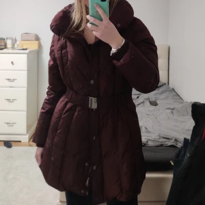 Dunjacka med indragen midja - Helt ny vinröd dunjacka med indragen midja och luva. Jackan är storlek 42/44 men passar även M och L. Den slutar mitten av låren, skriv för fler bilder!