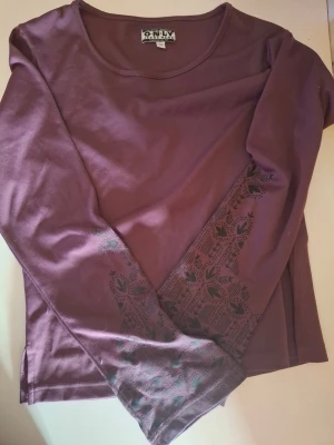 Lila långärmad topp från Only - Säljer en lila långärmad topp från Only med rund halsringning. Toppen har ett diskret svart mönster med blommor och blad på ärmarna, vilket ger en unik touch. Tillverkad i mjuk bomull som känns skön mot huden. Perfekt för dig som gillar stilrena men ändå personliga plagg.