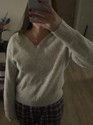 Beige ribbstickad tröja med v-ringning - Mysig beige ribbstickad tröja med bred v-ringning och långa ärmar. Tröjan har en avslappnad passform och breda muddar vid ärmslut och nederkant. Perfekt för lager på lager och enkel att matcha med olika stilar.