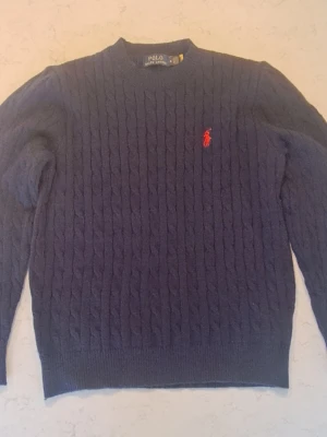 Mörkblå kabelstickad tröja Polo Ralph Lauren - Mörkblå kabelstickad tröja från Polo Ralph Lauren med rund halsringning och röd broderad logga på bröstet. Tröjan har långa ärmar och ribbade muddar vid ärmslut och nederkant. Perfekt för en stilren och klassisk look.