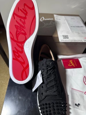 Christian Louboutin svarta sneakers - Svarta sneakers från Christian Louboutin med klassisk röd sula och svarta nitar på tån. Skorna har en låg siluett, svarta skosnören och är tillverkade i textil och läder. Perfekt för dig som vill sticka ut med exklusiva detaljer och edgy vibe.