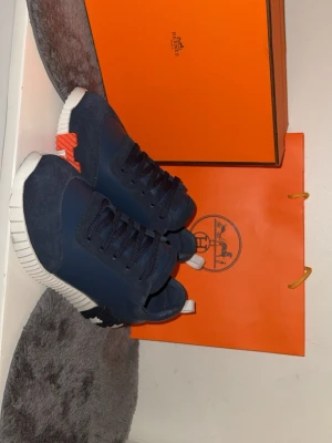 Mörkblå H-Sneakers - Snygga mörkblå sneakers med vit och svart sula samt orange detalj vid hälen. Skorna har en mix av mocka och slätt läder, samt klassiska snörningar. Ikonisk H-detalj på sidan och modern, chunky sula för en trendig look.