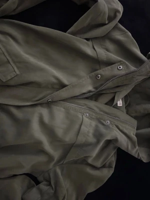 Olivgrön vindjacka från Jacqueline de Yong - Snygg olivgrön vindjacka från Jacqueline de Yong med dragkedja och tryckknappar framtill. Jackan har huva och en ficka på sidan. Perfekt för blåsiga dagar och enkel att matcha med olika outfits.