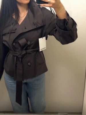 Kort brun trenchcoat från BERSHKA  - Snygg kort trenchcoat i mörkbrunt från Bershka. Jackan har dubbelknäppning, bred krage, axelklaffar och knytskärp i midjan. Tillverkad i ett kraftigt tyg med detaljerade manschetter och fickor framtill. Jackan är helt ny och prislappen sitter kvar, jackan är helt slutsåld på hemsidan