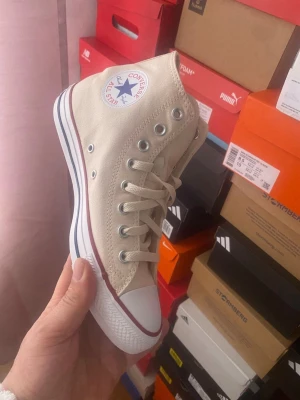 Beige Converse Chuck Taylor High - Snygga beige Converse Chuck Taylor All Star high tops i canvas med klassisk vit tåhätta i gummi, vita snören och metallfärgade öljetter. Ikonisk logga på sidan och randig sula i vitt med röda och svarta detaljer. Perfekta för en clean och stilren look.
