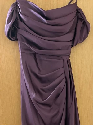 Lång lila klänning med drapering - Elegant långklänning i lila satin med draperad överdel och puffiga korta ärmar. Klänningen har en axelbandsdetalj och vacker fallande kjol. Perfekt för speciella tillfällen där du vill sticka ut med en unik och stilren look.