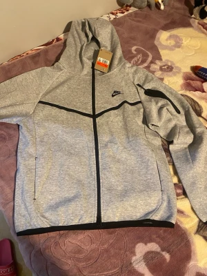 Gråa Nike Tech Fleece hoodie och byxor  - Snygga gråa Nike Tech Fleece mjukisbyxor och hoodie med svarta dragkedjor och logga på benet. Byxorna har smal passform och är tillverkade i mjukt och stretchigt material som ger en sportig look. Perfekta för chill eller träning.