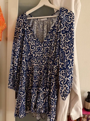 Blåvit mönstrad kortklänning Zara - Säljer en blåvit kortklänning från Zara med v-ringning och långa ärmar. Klänningen har ett unikt abstrakt mönster och är tillverkad i ett lätt material som passar perfekt för varmare dagar. Flowy passform och snygg detalj med knyt i nacken.
