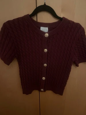 Vinröd kabelstickad kofta kort ärm - Kortärmad vinröd kofta med kabelstickat mönster och dekorativa guldknappar framtill. Rund halsringning och ribbade kanter ger en snygg och trendig look. Perfekt att styla över ett linne eller t-shirt för en chill vibe.