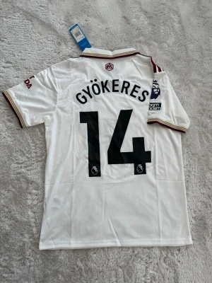 Arsenal vit matchtröja Adidas - Snygg vit Arsenal matchtröja från Adidas med vinröda detaljer och klubbmärke på bröstet. Tröjan har trycket 'GYÖKERES 14' på ryggen och Premier League-märke på ärmen. Tillverkad i lätt och ventilerande material, perfekt för fotboll eller träning.