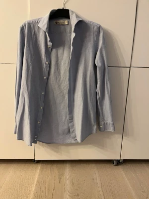 Ljusblå skjorta från Morris - Klassisk ljusblå skjorta från Morris i bomull. Skjortan har lång ärm, vit knappstängning framtill och enkel krage. Perfekt till både jeans och chinos för en clean och stilren look. Storlek M