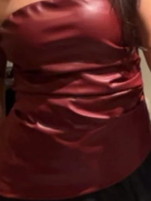 Vinröd tubtopp i skinnimitation - Säljer en vinröd axelbandslös tubtopp i skinnimitation. Toppen har en glansig finish och sitter tight mot kroppen. Perfekt för dig som vill sticka ut med en edgy och trendig look.