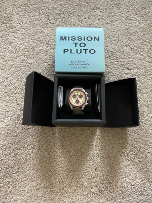 Omega x Swatch Mission to Pluto klocka - Säljer en Omega x Swatch Mission to Pluto från Bioceramic MoonSwatch Collection. Klockan har en beige urtavla med vinröda detaljer och en vinröd bezel. Armbandet är grönt och klockan är tillverkad i biokeramik. Perfekt för dig som gillar unika accessoarer!