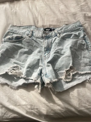 Ljusa denimshorts med slitningar - Säljer ett par ljusblå denimshorts från 157 med råa kanter och slitna detaljer framtill. Klassisk femficksmodell med knapp och dragkedja. Perfekta för varma dagar och ger en avslappnad look.