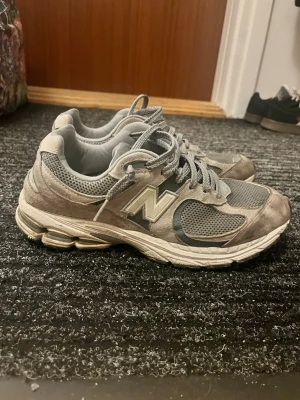 Grå New Balance 2002R sneakers - Säljer ett par grå New Balance 2002R sneakers med mesh och mockadetaljer. Skorna har klassisk N-logga på sidan, tjock sula och snörning. Perfekta för dig som gillar retrostil och bekväma skor till vardags. Färgskalan går i olika nyanser av grått.