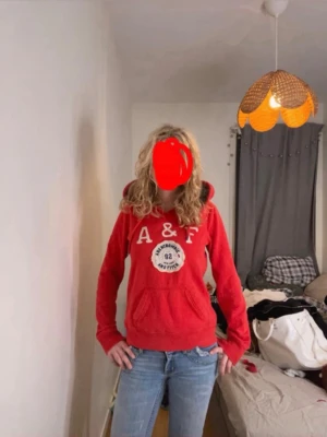 Röd hoodie från Abercrombie & Fitch - Snygg röd hoodie från Abercrombie & Fitch med broderad logga och text på bröstet. Mjuk insida och klassisk känguruficka framtill. Dragsko i huvan och ribbade muddar vid ärmar och nederkant. Perfekt för en avslappnad och trendig look.